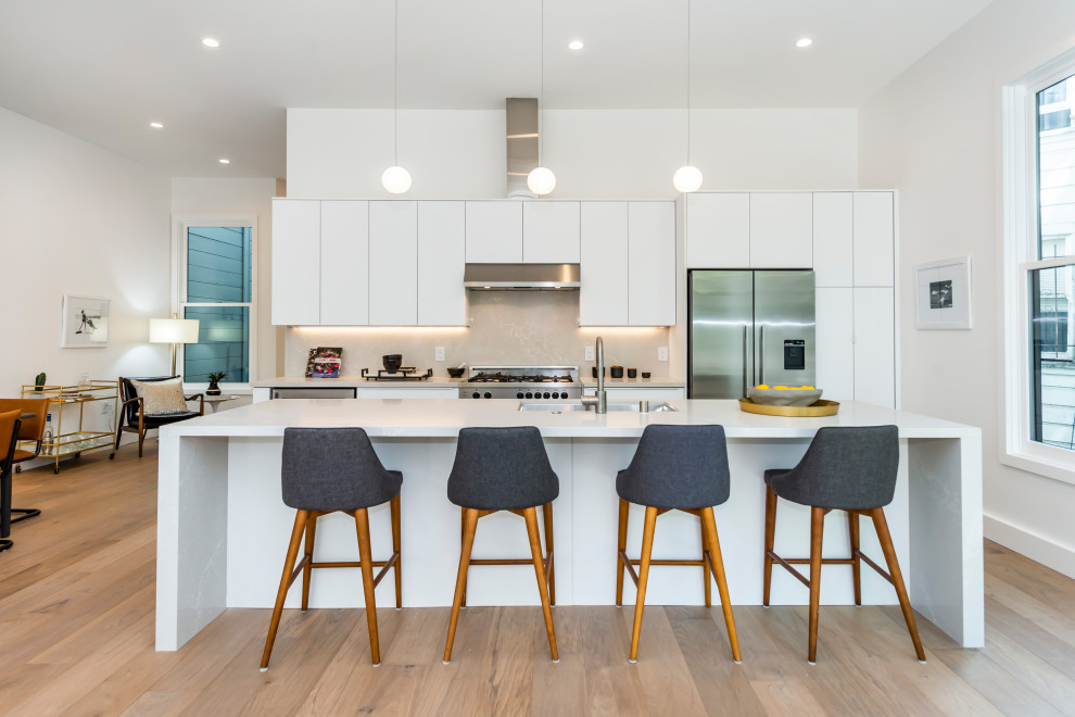 This is an example of a mid-sized contemporary galley open plan kitchen in San Francisco with an undermount sink, flat-panel cabinets, white cabinets, quartz benchtops, white splashback, engineered quartz splashback, stainless steel appliances, light hardwood floors, with island, beige floor and white benchtop.