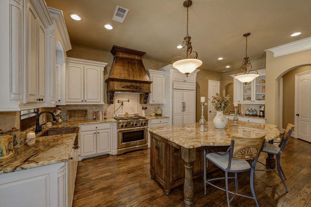 This is an example of a large traditional l-shaped open plan kitchen in Dallas with a farmhouse sink, distressed cabinets, granite benchtops, beige splashback, stone tile splashback, panelled appliances, medium hardwood floors, with island, raised-panel cabinets and brown floor.