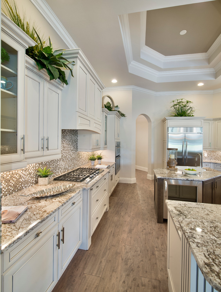 Photo of an expansive transitional separate kitchen in Miami with beaded inset cabinets, white cabinets, granite benchtops, mosaic tile splashback, stainless steel appliances, medium hardwood floors, multiple islands and brown floor.