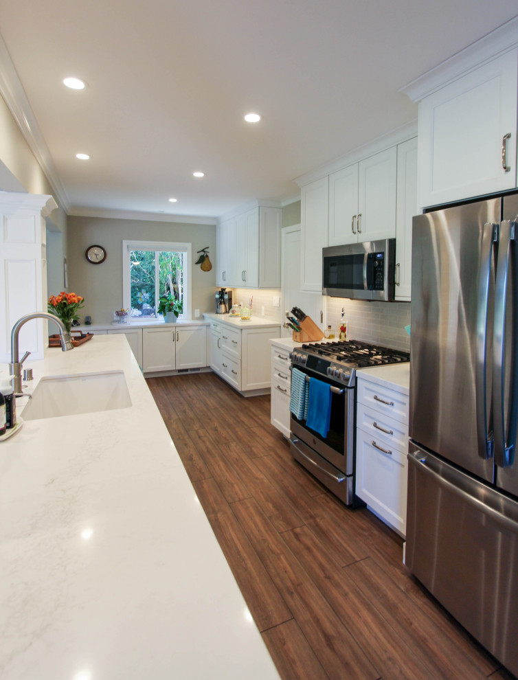 This is an example of a large traditional galley open plan kitchen in San Francisco with a submerged sink, recessed-panel cabinets, white cabinets, engineered stone countertops, grey splashback, metro tiled splashback, stainless steel appliances, vinyl flooring, an island, brown floors and white worktops.