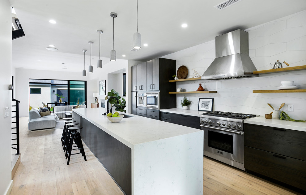 Inspiration for an expansive contemporary galley kitchen in Los Angeles with flat-panel cabinets, quartz benchtops, white splashback, porcelain splashback, stainless steel appliances, light hardwood floors, with island, white benchtop, an undermount sink, black cabinets and beige floor.
