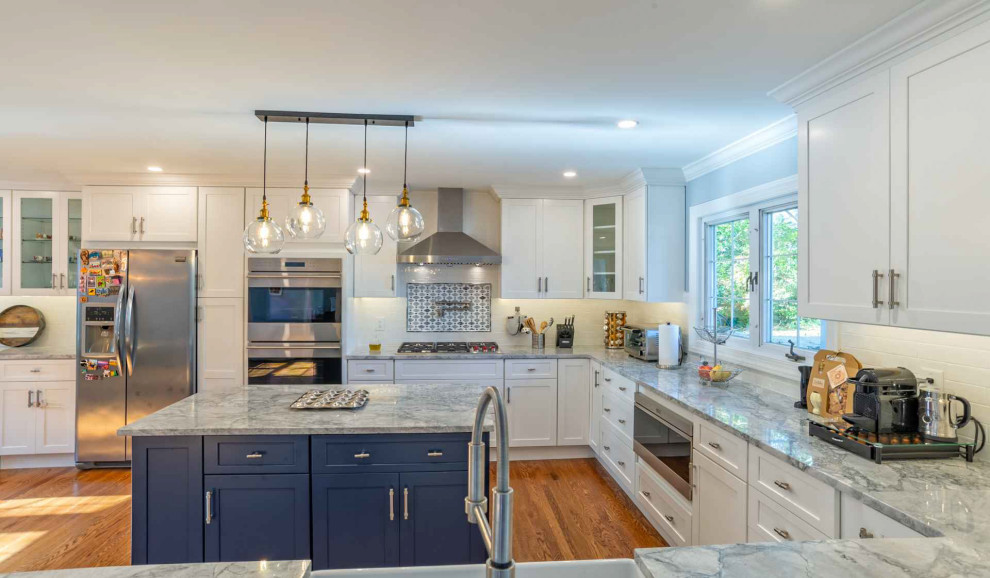 Photo of a mid-sized transitional u-shaped eat-in kitchen in Philadelphia with a farmhouse sink, shaker cabinets, blue cabinets, quartz benchtops, white splashback, porcelain splashback, stainless steel appliances, medium hardwood floors, with island, brown floor and grey benchtop.