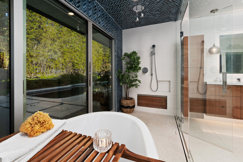 Пример оригинального дизайна: огромная главная ванная комната в стиле ретро с плоскими фасадами, фасадами цвета дерева среднего тона, отдельно стоящей ванной, душевой комнатой, унитазом-моноблоком, синей плиткой, стеклянной плиткой, синими стенами, полом из терраццо, настольной раковиной, стеклянной столешницей, бежевым полом, душем с распашными дверями, белой столешницей, тумбой под одну раковину и подвесной тумбой