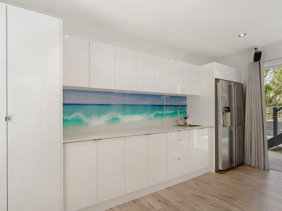 This is an example of a small contemporary single-wall open plan kitchen in Gold Coast - Tweed with a single-bowl sink, flat-panel cabinets, white cabinets, granite benchtops, blue splashback, glass sheet splashback, stainless steel appliances and light hardwood floors.