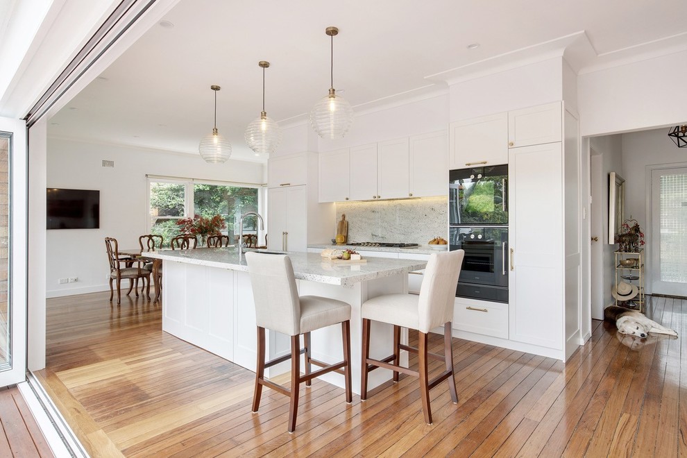 Design ideas for a mid-sized transitional single-wall open plan kitchen in Sydney with shaker cabinets, white cabinets, granite benchtops, grey splashback, marble splashback, black appliances, medium hardwood floors, with island, brown floor and white benchtop.