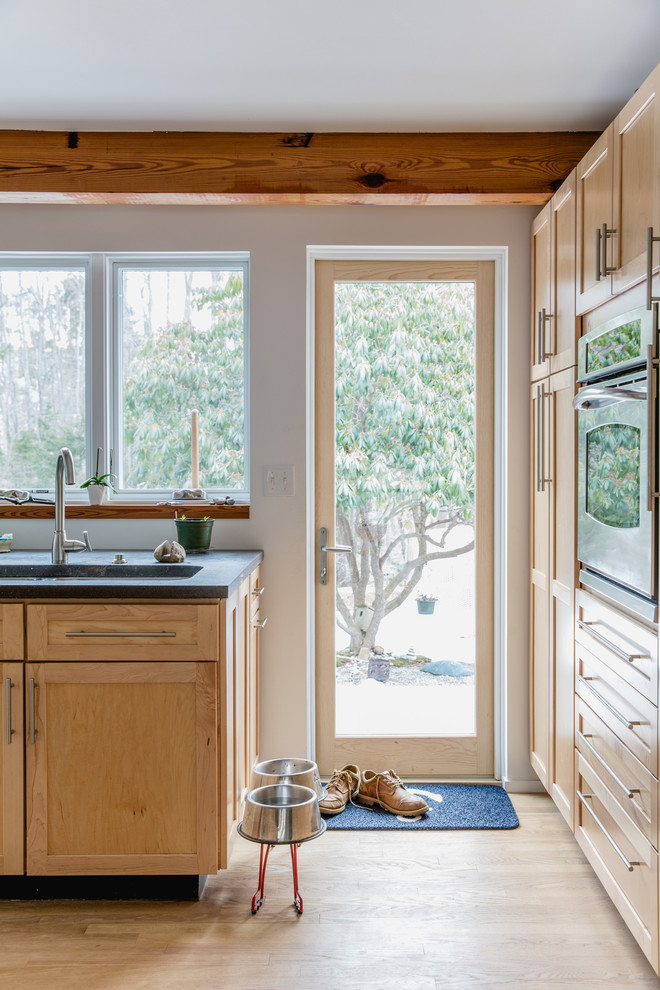 Photo of a country kitchen in Portland Maine with an undermount sink, shaker cabinets, light wood cabinets, soapstone benchtops, stainless steel appliances, light hardwood floors and beige floor.