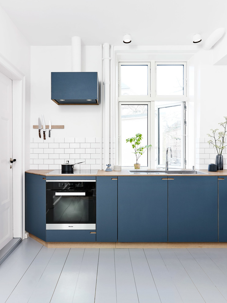 Photo of a mid-sized scandinavian l-shaped eat-in kitchen in Copenhagen with a drop-in sink, beaded inset cabinets, blue cabinets, wood benchtops, white splashback, ceramic splashback, stainless steel appliances, painted wood floors, a peninsula and white floor.