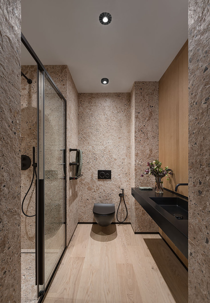 Minimalistisk inredning av ett mellanstort svart svart toalett, med släta luckor, svarta skåp, en toalettstol med hel cisternkåpa, beige kakel, keramikplattor, beige väggar, mellanmörkt trägolv, ett väggmonterat handfat, bänkskiva i akrylsten och beiget golv