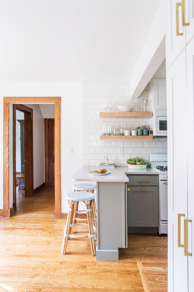 Example of a small transitional u-shaped medium tone wood floor and brown floor eat-in kitchen design with a farmhouse sink, shaker cabinets, blue cabinets, quartz countertops, white backsplash, porcelain backsplash, white appliances and a peninsula