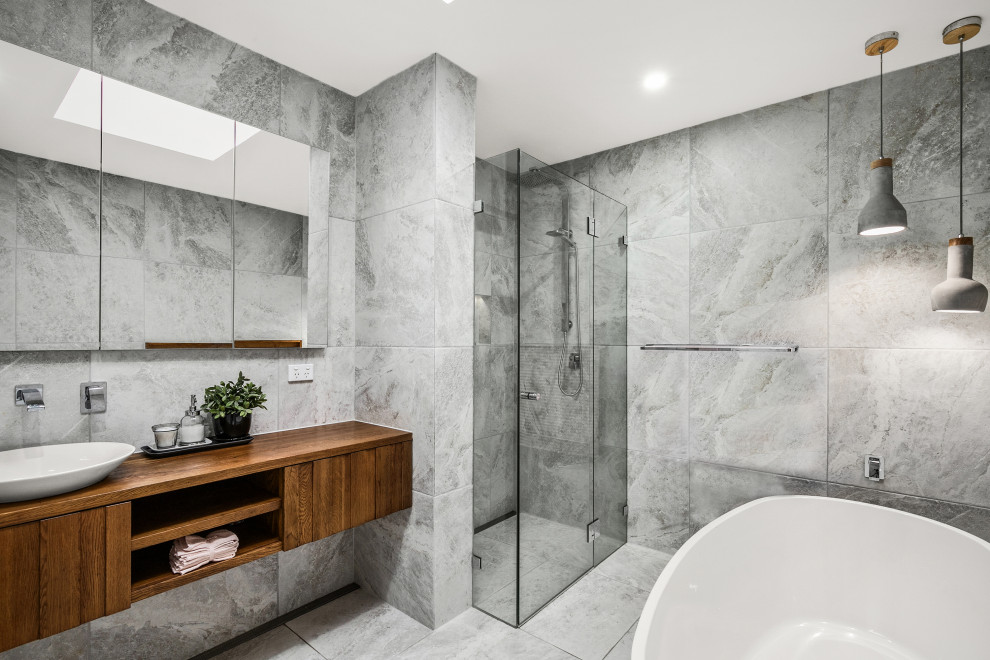 This is an example of a large contemporary grey and brown ensuite bathroom in Sydney with freestanding cabinets, medium wood cabinets, a freestanding bath, a corner shower, a one-piece toilet, grey tiles, ceramic tiles, grey walls, a vessel sink, wooden worktops, grey floors, a hinged door, brown worktops, a single sink, a floating vanity unit, ceramic flooring and feature lighting.