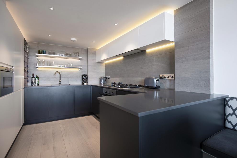 Photo of a small industrial u-shaped open plan kitchen in London with a drop-in sink, flat-panel cabinets, grey cabinets, quartz benchtops, grey splashback, ceramic splashback, light hardwood floors and a peninsula.