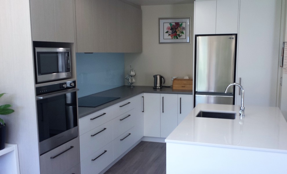 This is an example of a mid-sized contemporary l-shaped open plan kitchen in Christchurch with a single-bowl sink, flat-panel cabinets, white cabinets, quartz benchtops, blue splashback, glass sheet splashback, stainless steel appliances, vinyl floors, with island, grey floor and white benchtop.