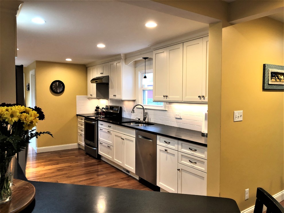 Example of a small transitional galley medium tone wood floor and brown floor enclosed kitchen design in Boston with an undermount sink, shaker cabinets, white cabinets, granite countertops, white backsplash, subway tile backsplash, stainless steel appliances and black countertops