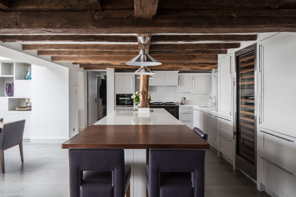 Farmhouse l-shaped open plan kitchen in Sussex with shaker cabinets, white cabinets, light hardwood flooring, an island and exposed beams.
