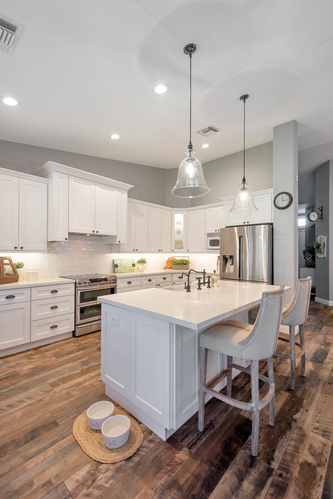 This is an example of a large country l-shaped open plan kitchen in Phoenix with a farmhouse sink, shaker cabinets, white cabinets, quartz benchtops, white splashback, subway tile splashback, stainless steel appliances, with island and white benchtop.
