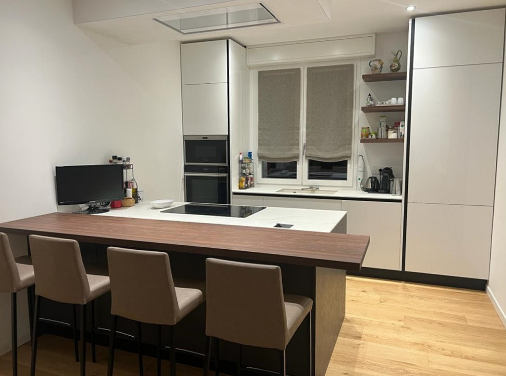 Inspiration for a medium sized contemporary galley open plan kitchen in Milan with a submerged sink, flat-panel cabinets, white cabinets, black appliances, light hardwood flooring, a breakfast bar, white worktops and a drop ceiling.