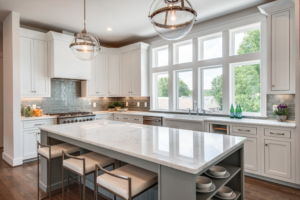 Inspiration for a mid-sized transitional l-shaped medium tone wood floor eat-in kitchen remodel in Nashville with a farmhouse sink, shaker cabinets, white cabinets, marble countertops, gray backsplash, glass tile backsplash, stainless steel appliances, an island and white countertops