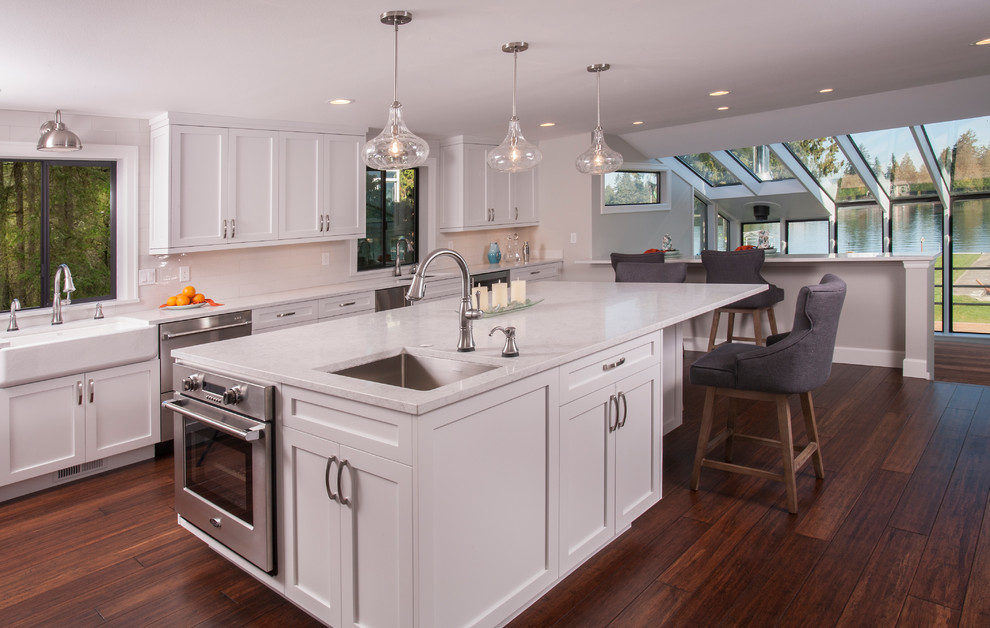 Large arts and crafts kitchen in Seattle with a farmhouse sink, shaker cabinets, white cabinets, quartz benchtops, grey splashback, porcelain splashback, stainless steel appliances, bamboo floors, with island, brown floor and white benchtop.