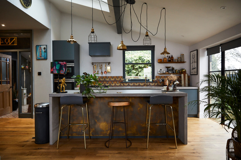 Design ideas for an eclectic galley kitchen in London with a submerged sink, flat-panel cabinets, blue cabinets, multi-coloured splashback, black appliances, medium hardwood flooring, an island, brown floors, grey worktops and a vaulted ceiling.