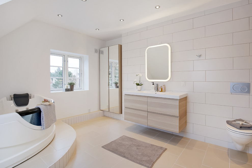 Inspiration for a large contemporary master bathroom in Copenhagen with flat-panel cabinets, beige cabinets, a corner tub, a wall-mount toilet, white tile, porcelain tile, white walls, an integrated sink and laminate benchtops.