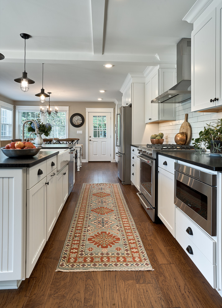Inspiration for a mid-sized country galley eat-in kitchen in Chicago with a farmhouse sink, shaker cabinets, white cabinets, granite benchtops, white splashback, ceramic splashback, stainless steel appliances, medium hardwood floors, with island, brown floor, black benchtop and coffered.