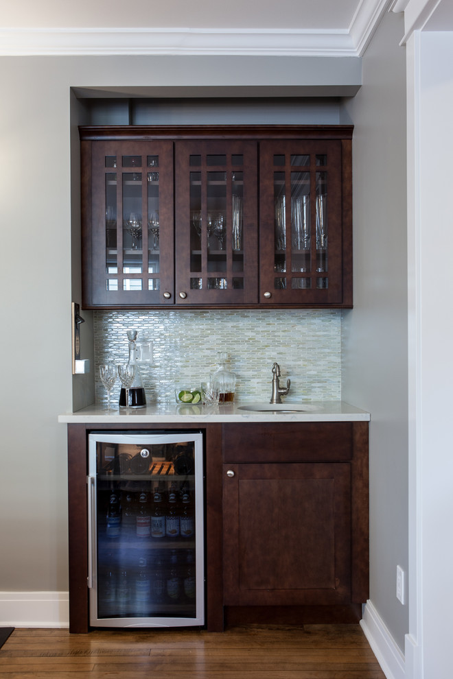 Photo of a small traditional single-wall wet bar in Chicago with an undermount sink, shaker cabinets, dark wood cabinets, quartz benchtops, grey splashback, glass tile splashback and medium hardwood floors.