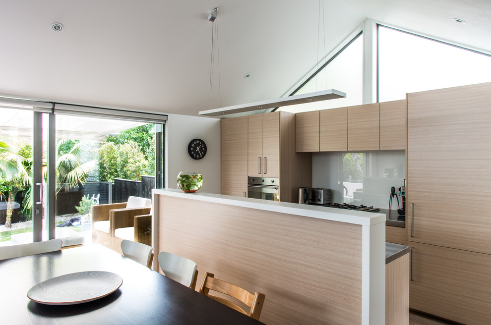 Design ideas for a small modern single-wall eat-in kitchen in Auckland with a drop-in sink, flat-panel cabinets, light wood cabinets, stainless steel benchtops, white splashback, glass sheet splashback, stainless steel appliances, light hardwood floors and with island.