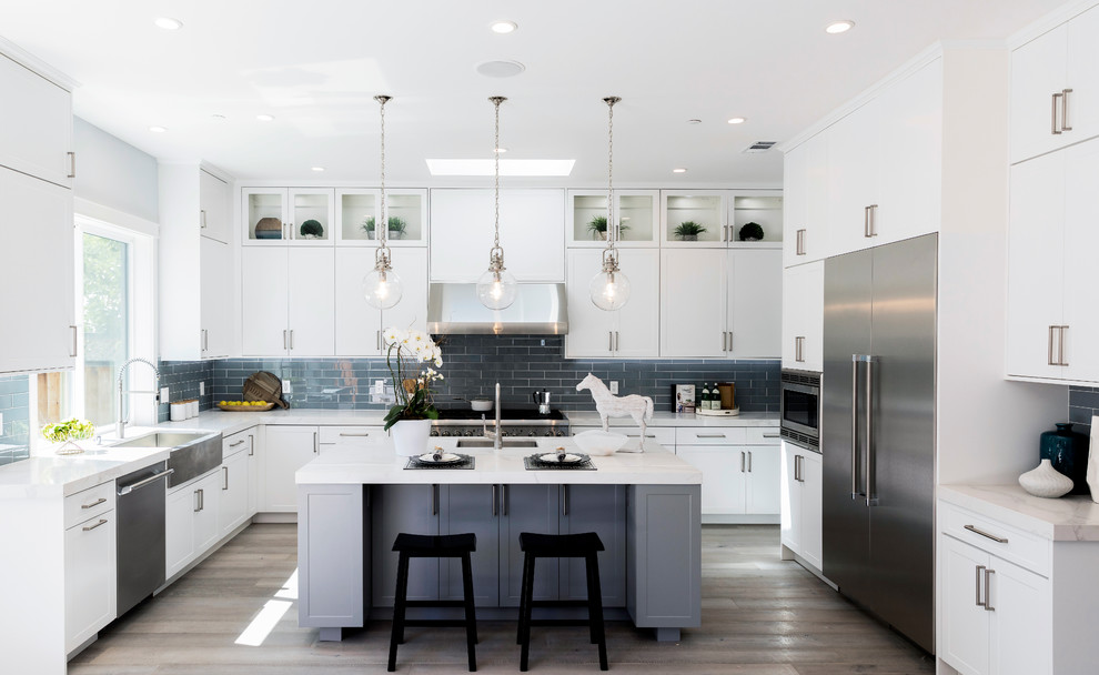 Photo of a transitional u-shaped kitchen in San Francisco with a farmhouse sink, shaker cabinets, white cabinets, subway tile splashback, stainless steel appliances, light hardwood floors, with island and white benchtop.