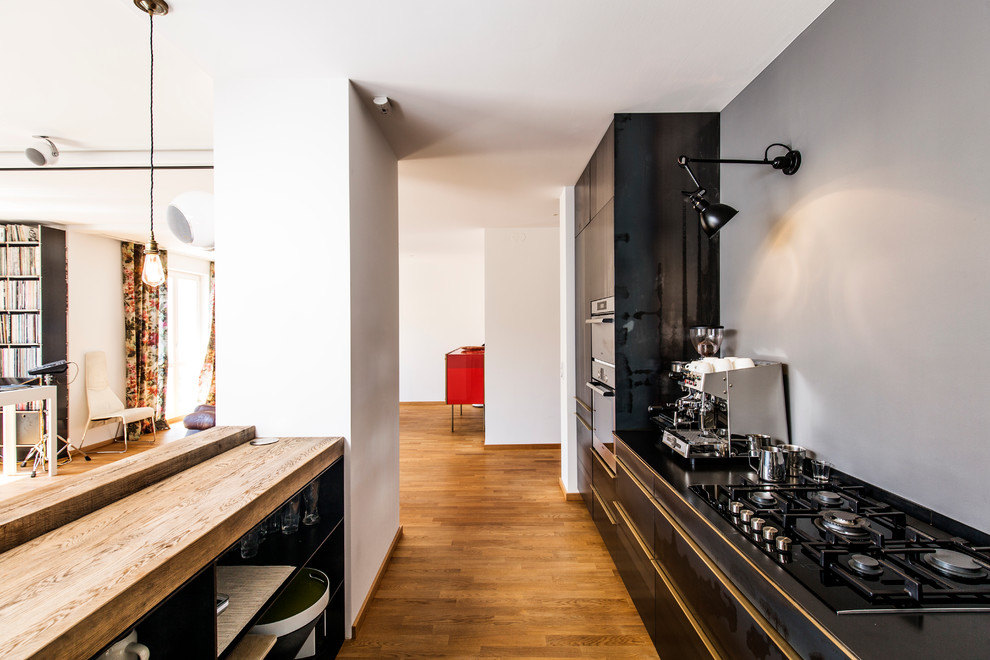 This is an example of a large contemporary galley open plan kitchen in Munich with flat-panel cabinets, black cabinets, medium hardwood floors, wood benchtops, grey splashback, black appliances, an integrated sink, no island and brown floor.