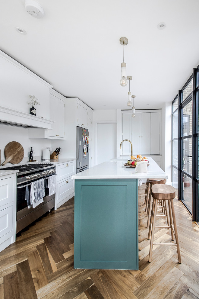 Inspiration for a mid-sized transitional galley open plan kitchen in Surrey with a farmhouse sink, shaker cabinets, white cabinets, quartz benchtops, white splashback, engineered quartz splashback, stainless steel appliances, medium hardwood floors, a peninsula, brown floor and white benchtop.