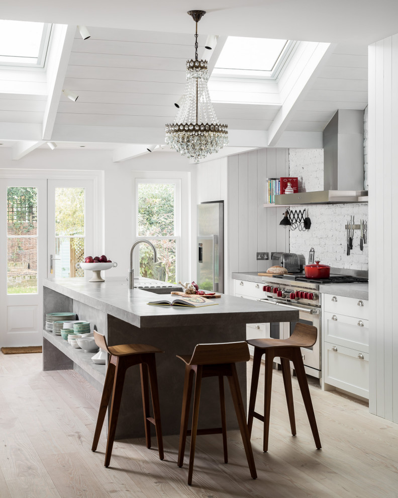 This is an example of a mid-sized country galley eat-in kitchen in London with an undermount sink, shaker cabinets, white cabinets, concrete benchtops, white splashback, brick splashback, stainless steel appliances, light hardwood floors, with island, beige floor and grey benchtop.