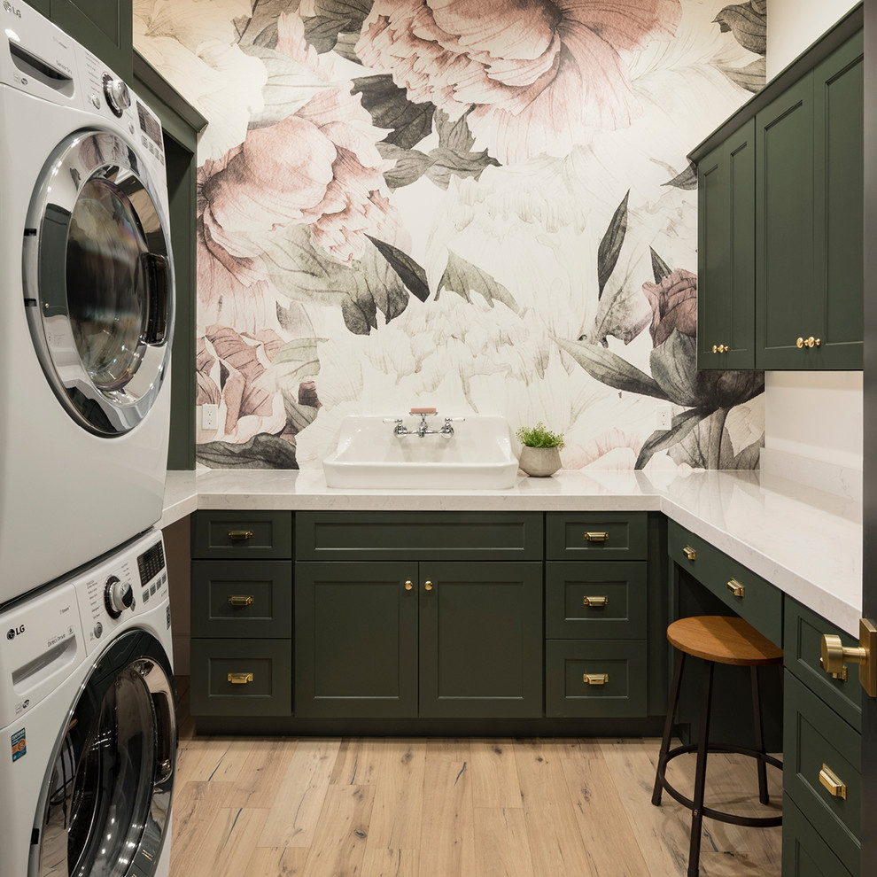 Photo of a transitional u-shaped dedicated laundry room in Phoenix with quartz benchtops, shaker cabinets, green cabinets, light hardwood floors, a stacked washer and dryer, brown floor, white benchtop and multi-coloured walls.