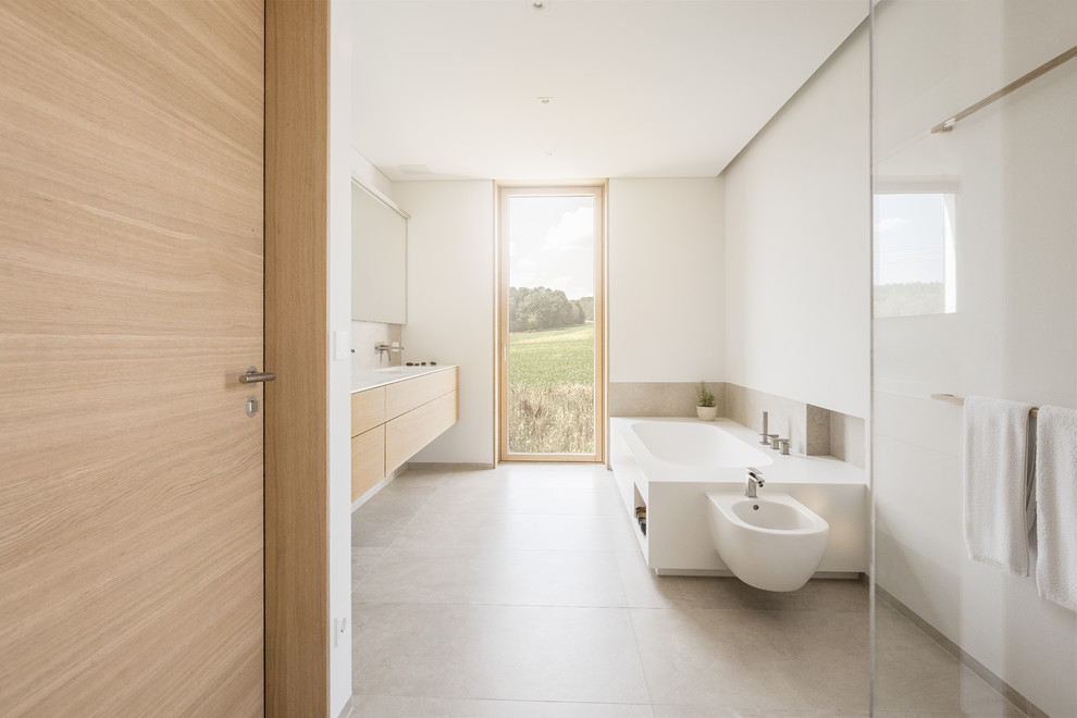This is an example of a large modern master bathroom in Nuremberg with flat-panel cabinets, medium wood cabinets, a drop-in tub, a curbless shower, a bidet, beige tile, ceramic tile, white walls, ceramic floors, a trough sink, solid surface benchtops, beige floor, an open shower and white benchtops.