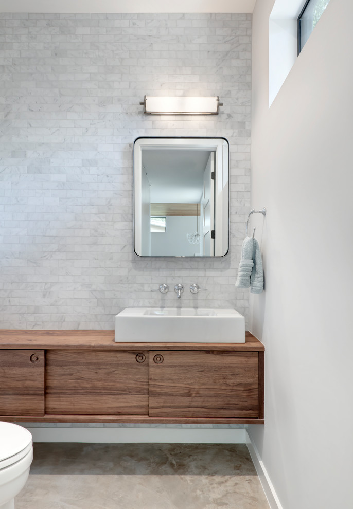 Photo of a mid-sized modern powder room in Austin with furniture-like cabinets, medium wood cabinets, a one-piece toilet, white tile, marble, white walls, concrete floors, a vessel sink, wood benchtops, grey floor and brown benchtops.