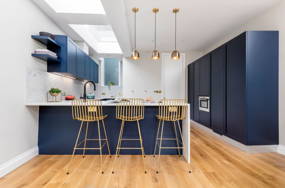 Design ideas for a mid-sized traditional l-shaped eat-in kitchen in London with an undermount sink, shaker cabinets, blue cabinets, quartzite benchtops, white splashback, engineered quartz splashback, panelled appliances, light hardwood floors, a peninsula, brown floor and yellow benchtop.