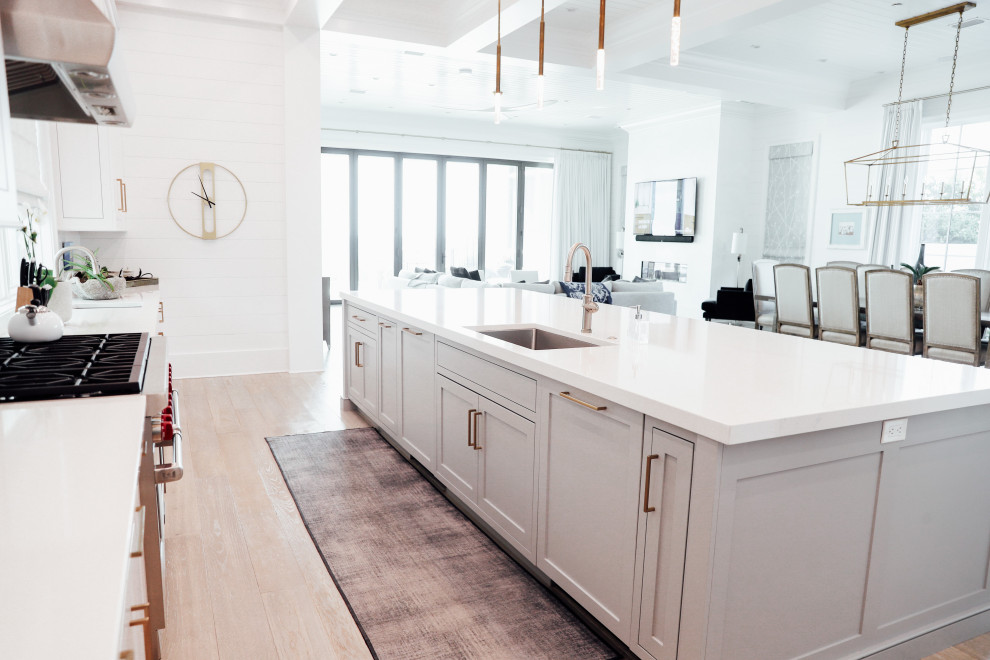 Maritim inredning av ett stort vit linjärt vitt kök och matrum, med en undermonterad diskho, skåp i shakerstil, bänkskiva i kvartsit, ljust trägolv, en köksö och brunt golv