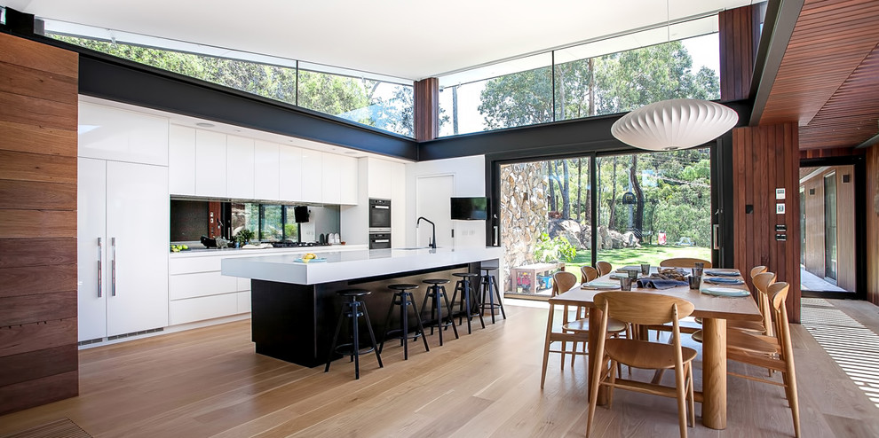 Inspiration for a mid-sized contemporary galley eat-in kitchen in Melbourne with an undermount sink, white cabinets, solid surface benchtops, mirror splashback, black appliances, medium hardwood floors and with island.