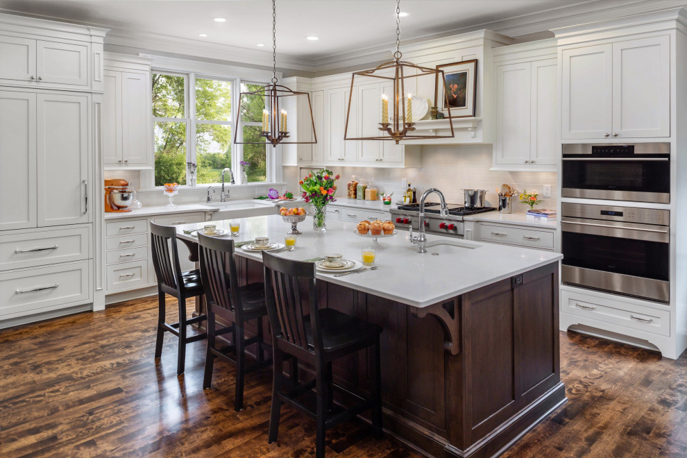 Traditional l-shaped kitchen in Milwaukee with a farmhouse sink, shaker cabinets, white cabinets, grey splashback, subway tile splashback, stainless steel appliances, dark hardwood floors, with island, brown floor and white benchtop.