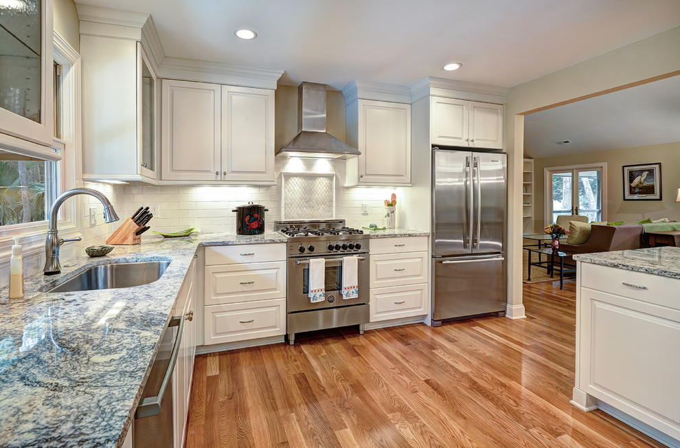 This is an example of a mid-sized transitional l-shaped kitchen in Charleston with a double-bowl sink, raised-panel cabinets, white cabinets, granite benchtops, white splashback, stainless steel appliances, light hardwood floors and with island.