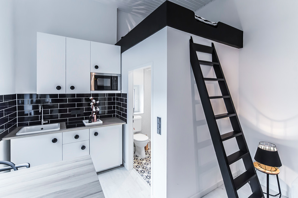 Photo of a small contemporary single-wall eat-in kitchen in Dusseldorf with a farmhouse sink, flat-panel cabinets, white cabinets, black splashback, subway tile splashback, stainless steel appliances, light hardwood floors, wood benchtops and a peninsula.