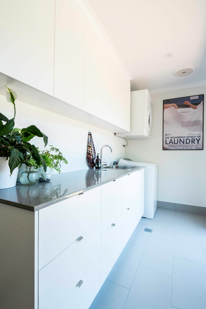 Photo of a large modern single-wall dedicated laundry room in Canberra - Queanbeyan with a drop-in sink, white cabinets, quartz benchtops, white walls, porcelain floors, a stacked washer and dryer and grey floor.
