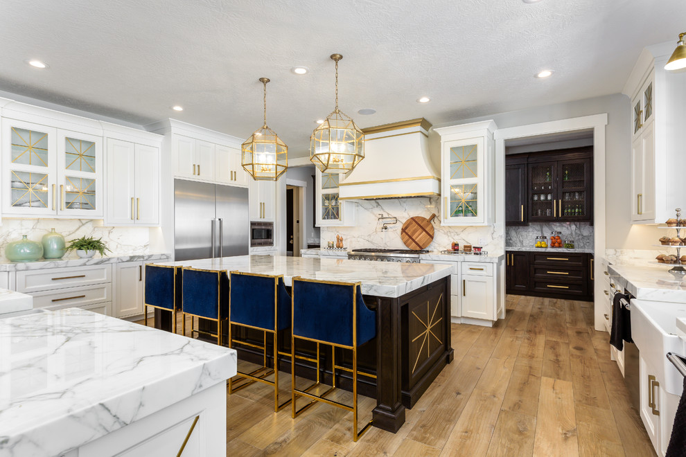 This is an example of a large transitional u-shaped open plan kitchen in Salt Lake City with a farmhouse sink, white cabinets, marble benchtops, white splashback, marble splashback, stainless steel appliances, light hardwood floors, multiple islands, white benchtop, glass-front cabinets and brown floor.