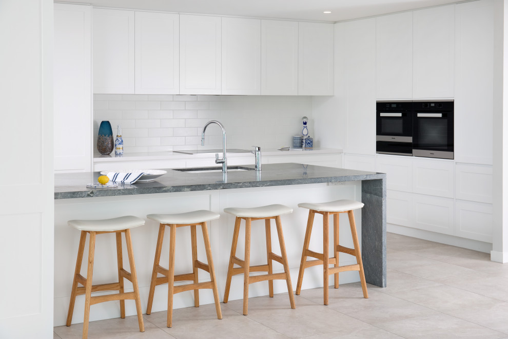 This is an example of a transitional u-shaped open plan kitchen in Brisbane with an undermount sink, shaker cabinets, white cabinets, white splashback, subway tile splashback, panelled appliances, with island, beige floor and grey benchtop.