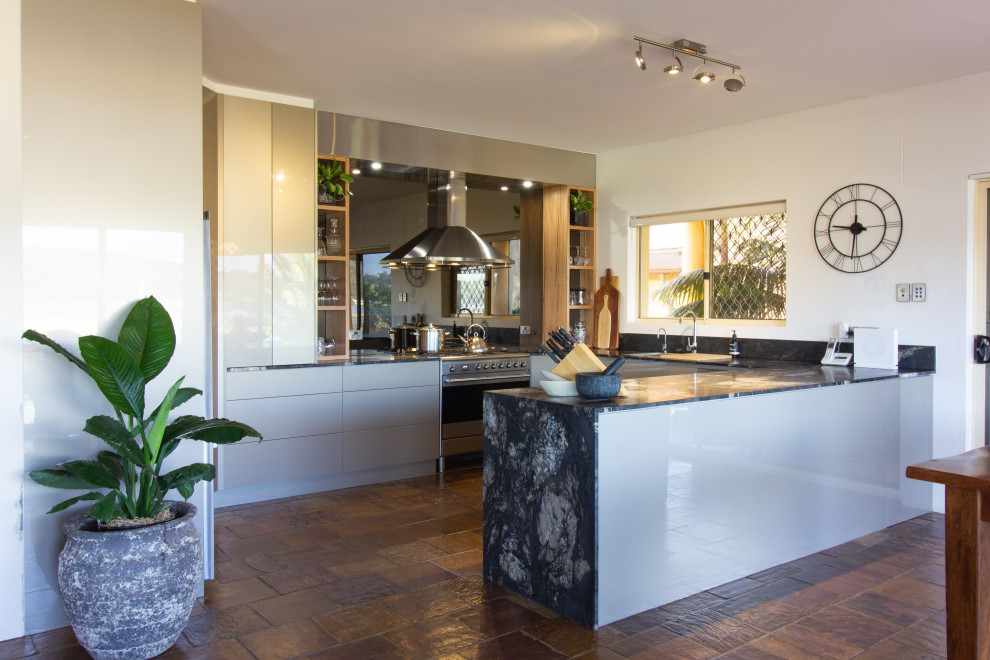 Large modern u-shaped kitchen pantry in Central Coast with a double-bowl sink, flat-panel cabinets, beige cabinets, granite benchtops, metallic splashback, glass sheet splashback, stainless steel appliances, slate floors, a peninsula, brown floor and black benchtop.