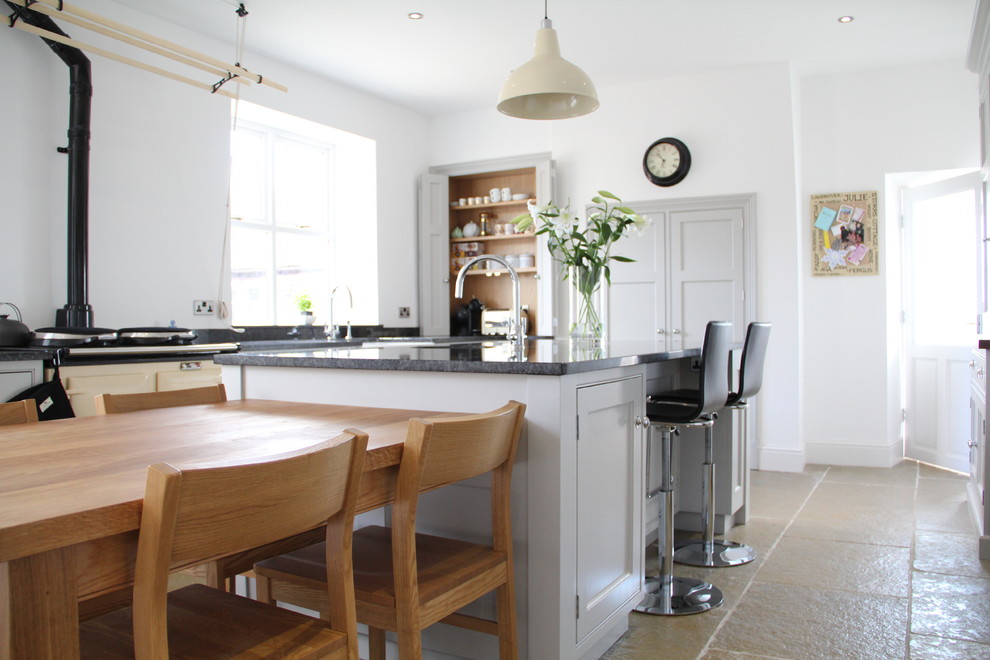 This is an example of a mid-sized traditional galley eat-in kitchen in Other with a farmhouse sink, recessed-panel cabinets, granite benchtops, stainless steel appliances, limestone floors, with island, grey cabinets, black splashback, beige floor and black benchtop.