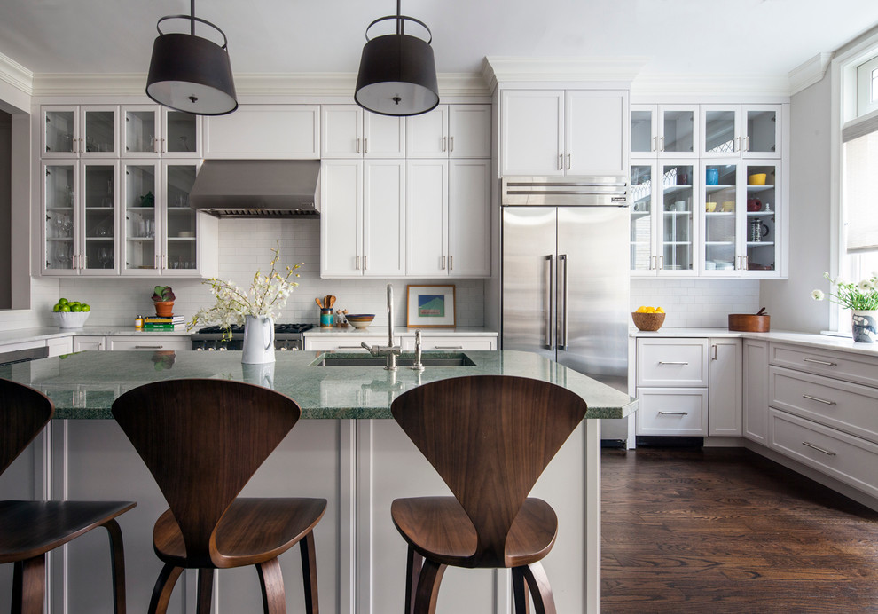This is an example of a large transitional u-shaped open plan kitchen in New York with glass-front cabinets, white cabinets, white splashback, subway tile splashback, stainless steel appliances, dark hardwood floors, with island, an undermount sink, marble benchtops and brown floor.