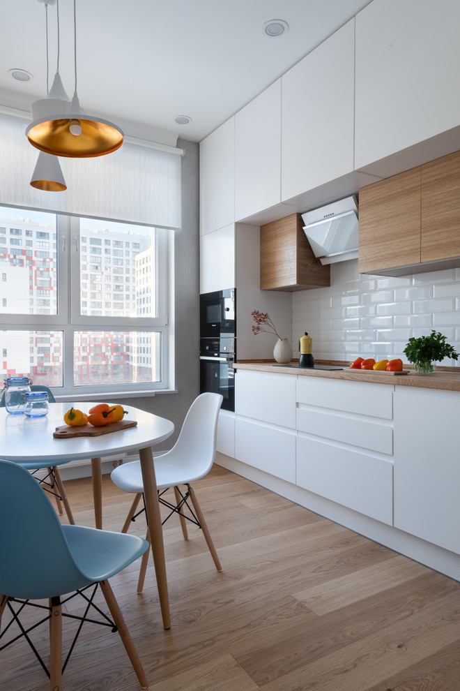 Design ideas for a contemporary eat-in kitchen in Moscow with flat-panel cabinets, white cabinets, wood benchtops, white splashback, subway tile splashback, black appliances, medium hardwood floors, brown floor, brown benchtop and no island.