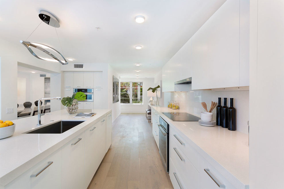 Photo of a mid-sized contemporary single-wall eat-in kitchen in Miami with an undermount sink, flat-panel cabinets, white cabinets, solid surface benchtops, white splashback, porcelain splashback, stainless steel appliances, light hardwood floors, with island, beige floor and white benchtop.