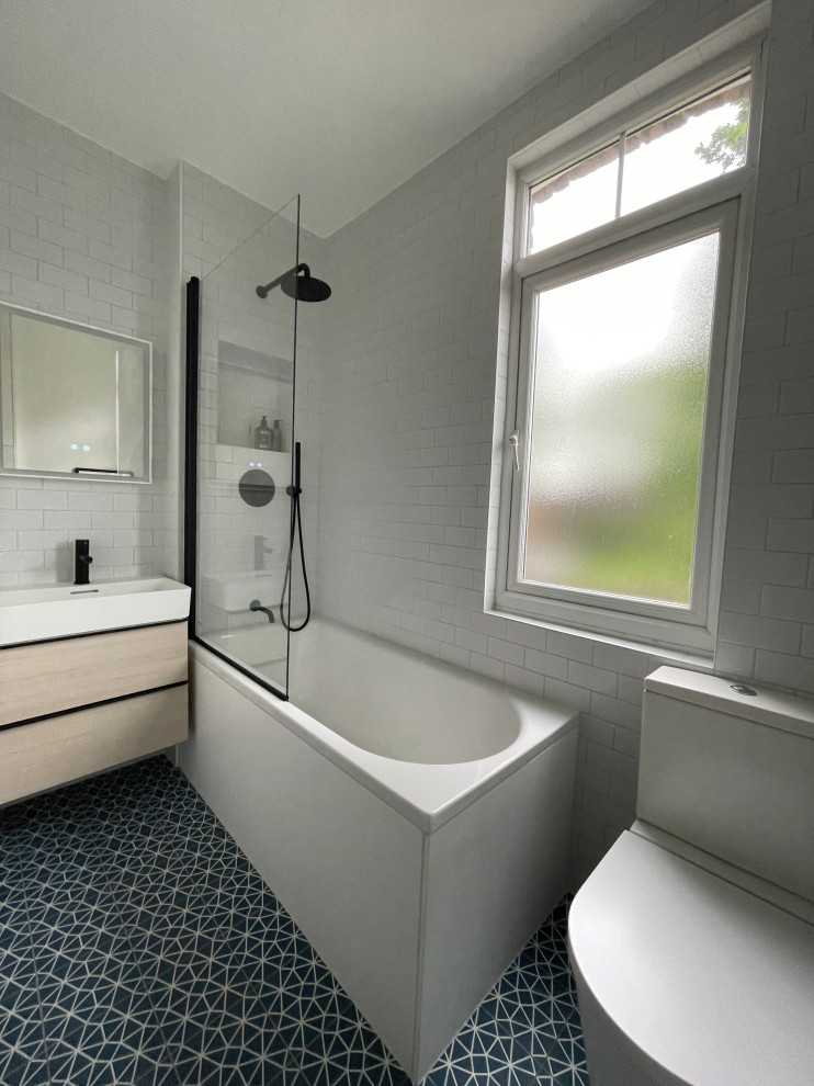 Photo of a small contemporary kids bathroom in London with flat-panel cabinets, light wood cabinets, a drop-in tub, a shower/bathtub combo, a two-piece toilet, white tile, porcelain tile, white walls, porcelain floors, a wall-mount sink, blue floor, a hinged shower door, a laundry, a single vanity and a floating vanity.