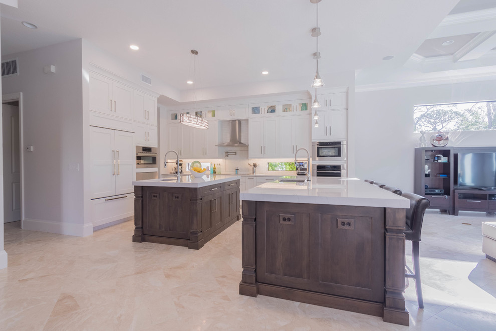 Open concept kitchen - huge transitional l-shaped porcelain tile and beige floor open concept kitchen idea with a farmhouse sink, shaker cabinets, white cabinets, quartz countertops, white backsplash, porcelain backsplash, stainless steel appliances and two islands